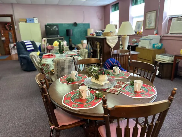 A round wooden table is set with Christmas placemats and mugs in the thrift shop furniture room.