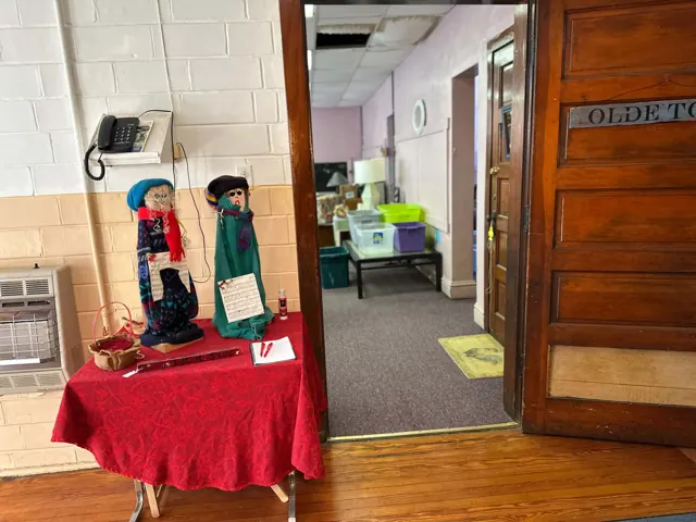 A decorated table stands by an open wooden doorway into a former classroom holding thrift shop furniture.