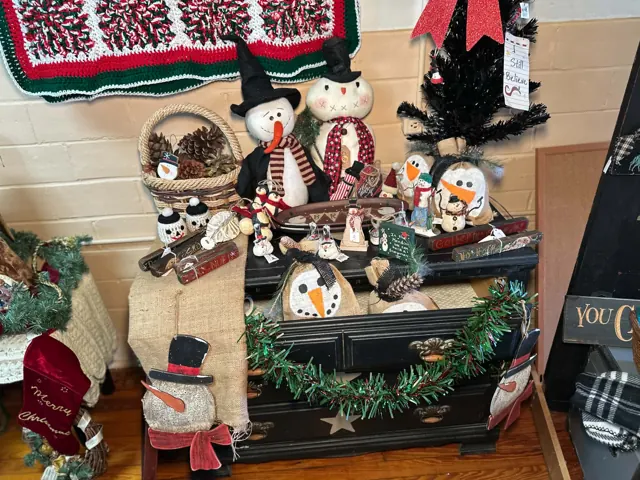 Snowmen, small holiday figures, baskets, and greenery are arranged on a dark wooden dresser.