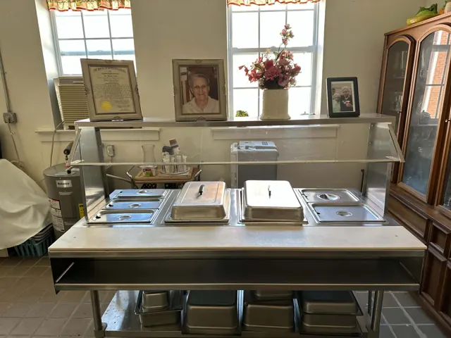 A stainless steel serving counter holds covered pans beneath framed photographs and a memorial tribute.
