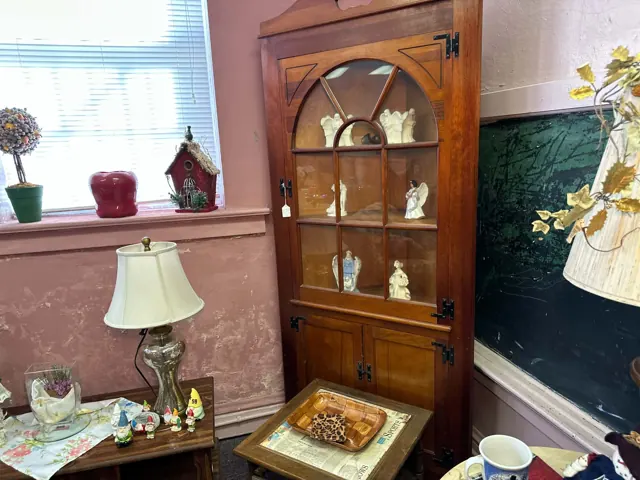 A tall wooden display cabinet holds small figures beside a lamp, side table, and classroom chalkboard.